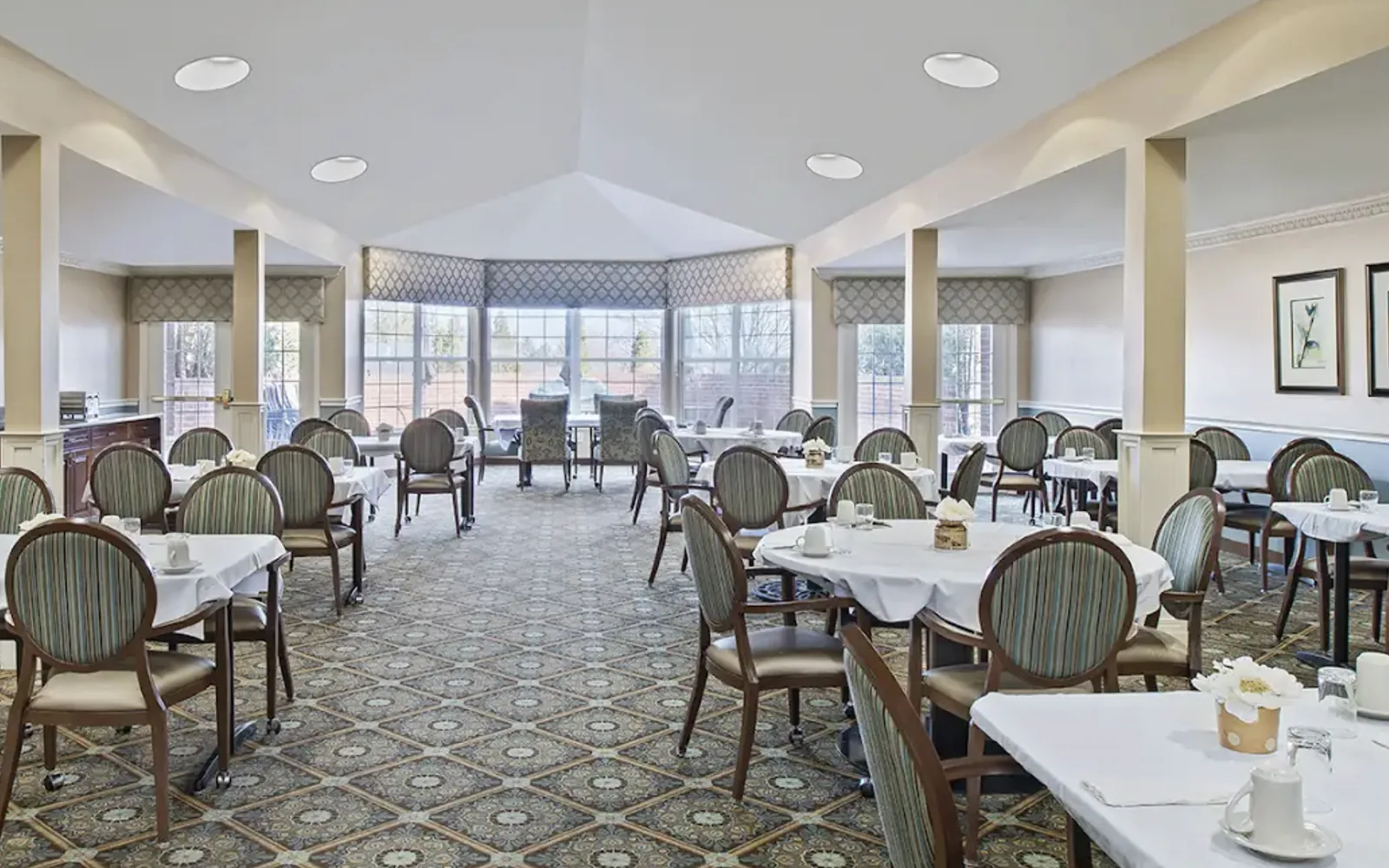 The Comfort Cove dining room, with round tables set in white linen beneath a bay of windows in West Bloomfield, MI