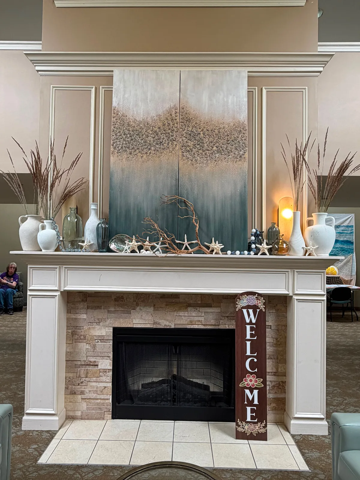 The lobby fireplace: a white mantel dressed with vases, starfish and driftwood beneath a coastal painting, and a wooden WELCOME sign at Comfort Cove Senior Living in West Bloomfield, MI