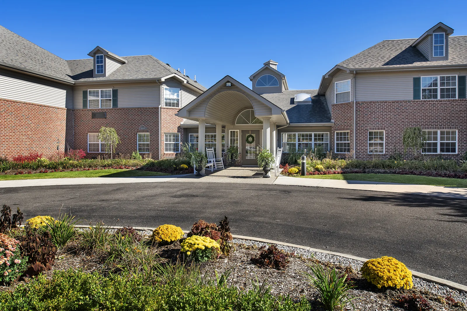 The front entrance of Comfort Cove: brick and siding building with a covered portico, rocking chairs and autumn planting in West Bloomfield, MI
