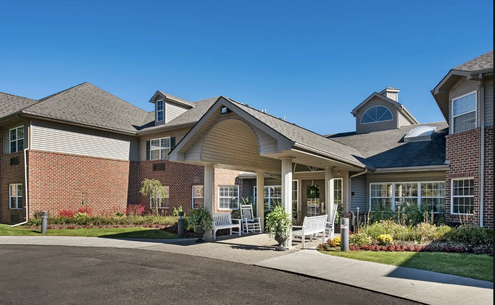 The covered front porch: rocking chairs and benches under the entrance portico, with flower beds along the drive at Comfort Cove Senior Living in West Bloomfield, MI