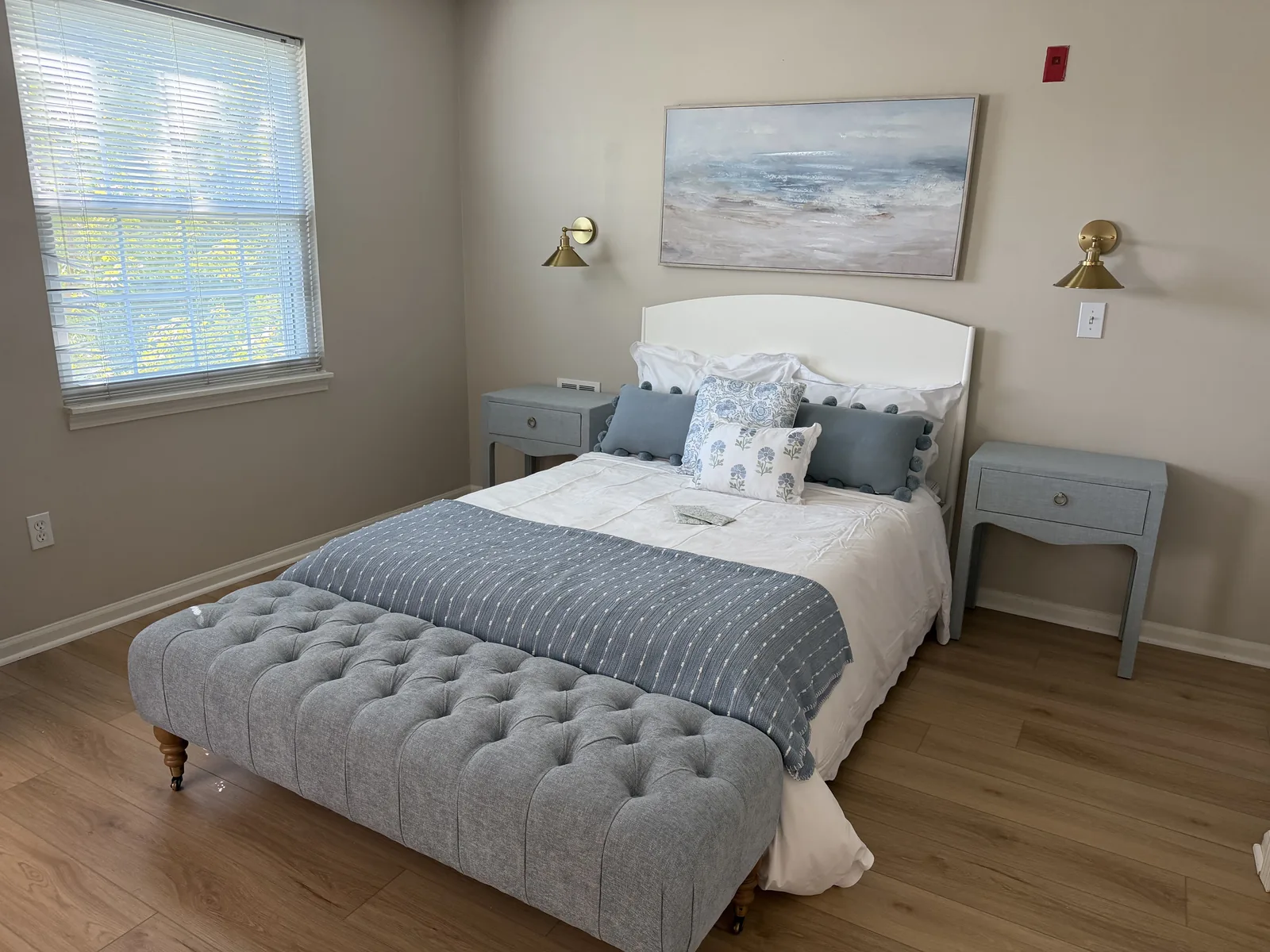 A one-bedroom suite: upholstered bed with blue and white linens between two nightstands, seascape above at Comfort Cove Senior Living in West Bloomfield, MI