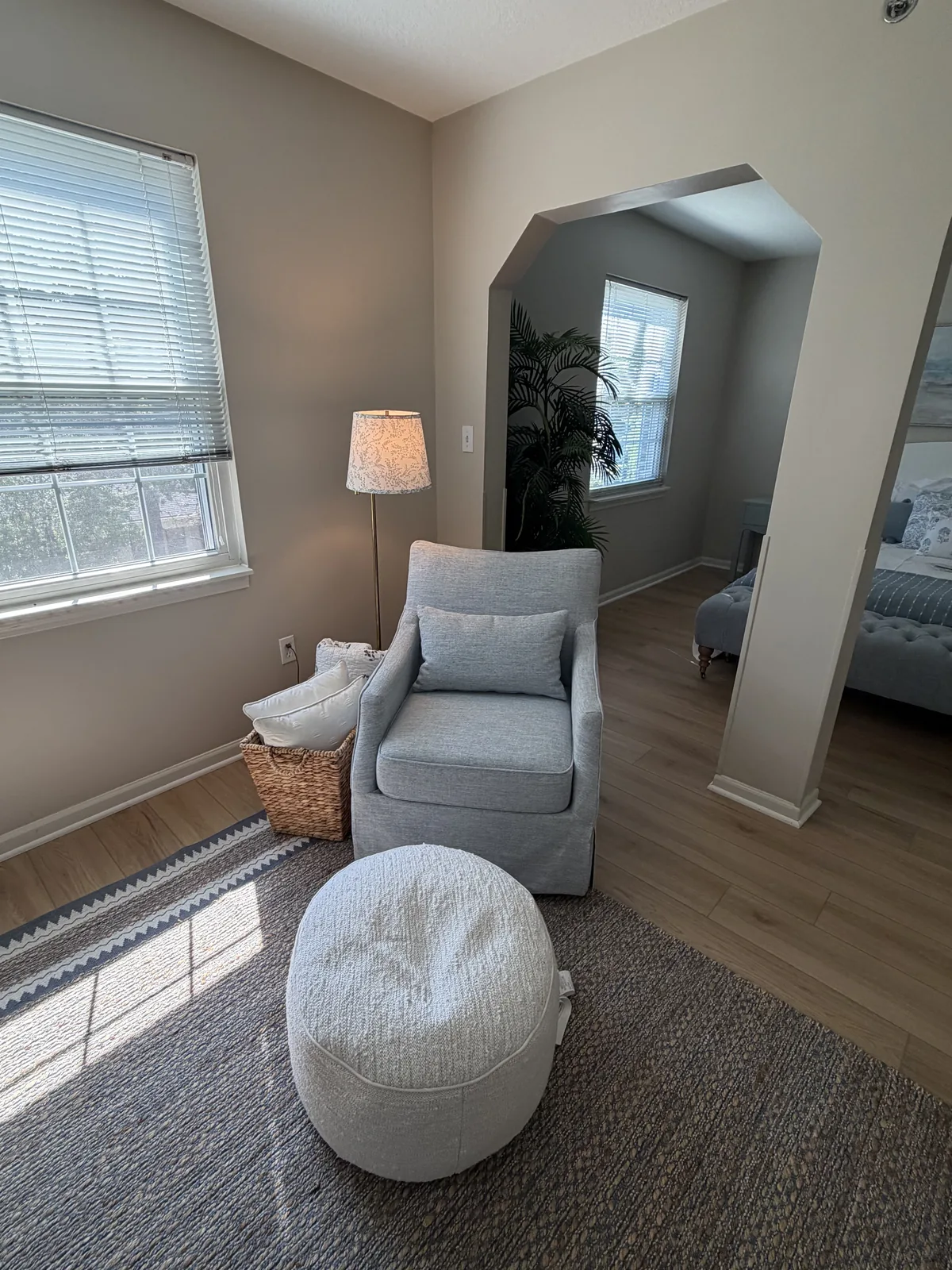An apartment living room: grey armchair and ottoman by the window, with a nook leading to the bedroom at Comfort Cove Senior Living in West Bloomfield, MI